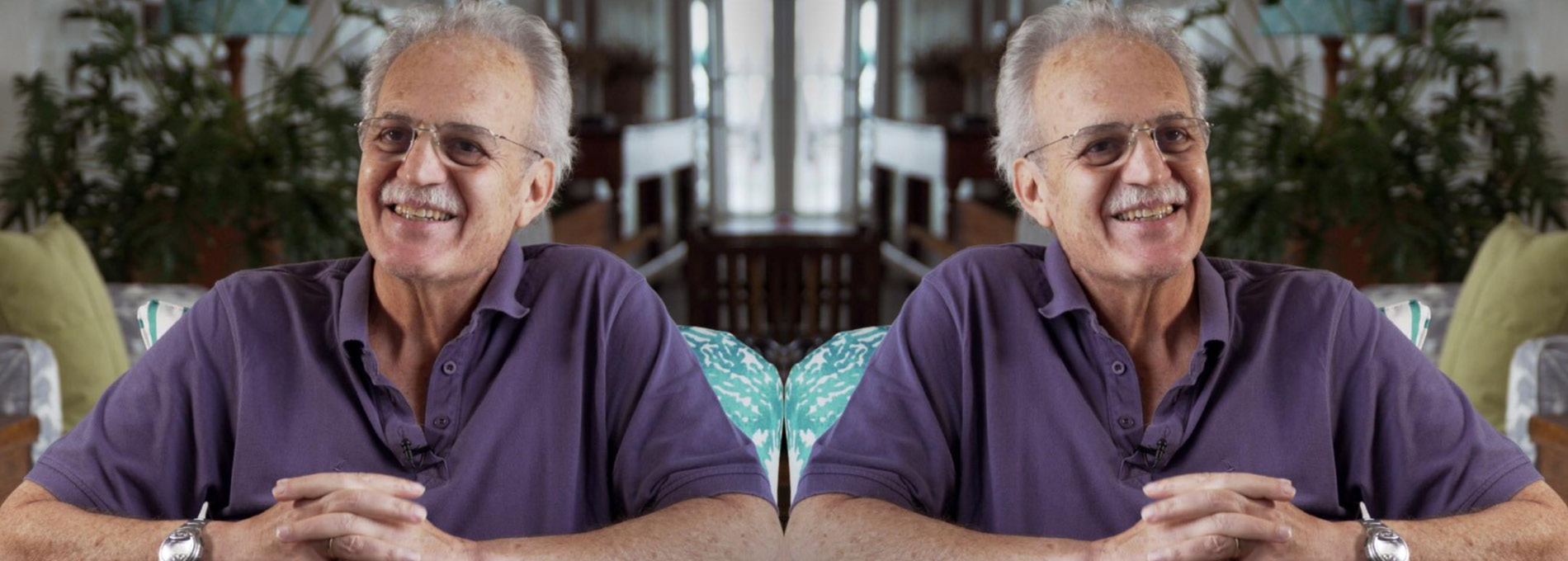 Carlos Nobre sua casa, sentado e apoiado em uma mesa, usando uma camisa gola polo roxa, óculos, relógio no pulso e sorrindo.