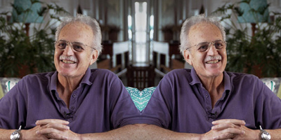 Carlos Nobre sua casa, sentado e apoiado em uma mesa, usando uma camisa gola polo roxa, óculos, relógio no pulso e sorrindo.